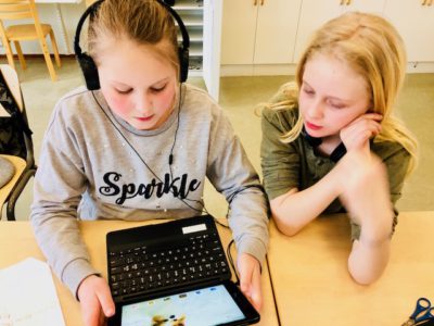 Två skolbarn sitter vid ett skrivbord med en dator. Ena barnet har på sig hörlurar.